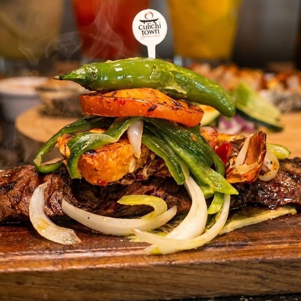 Steaming grilled steak with onions, peppers, and a Culiichi Town pick on a wooden board.