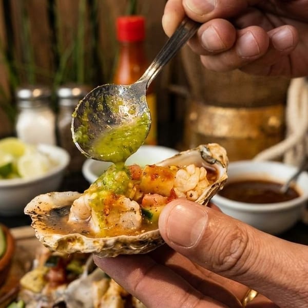 A hand holds an oyster shell filled with seafood and salsa, drizzling green sauce from a spoon.