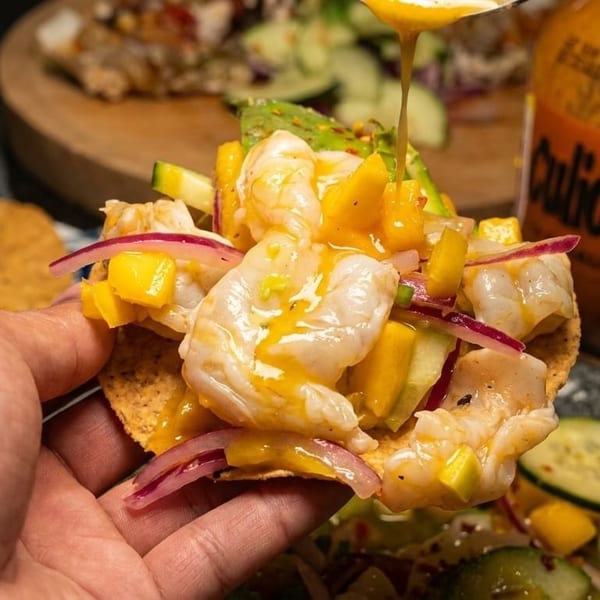 A hand holds a shrimp tostada topped with mango, red onion, and sauce drizzling from above.