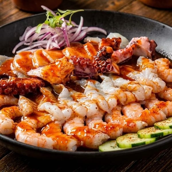 A black bowl filled with shrimp, octopus, and fish ceviche, garnished with red onion and cucumber.