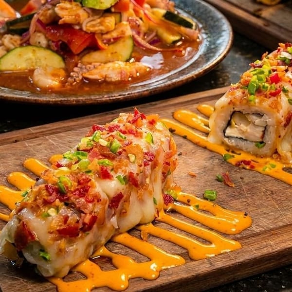 Two sushi rolls topped with bacon and green onions, drizzled with orange sauce, beside a ceviche dish.