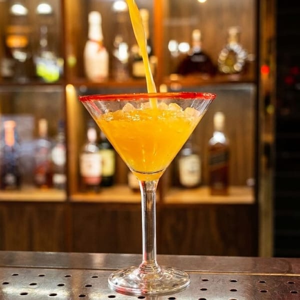 A bright orange cocktail with a red sugar rim is being poured into a martini glass at a bar.