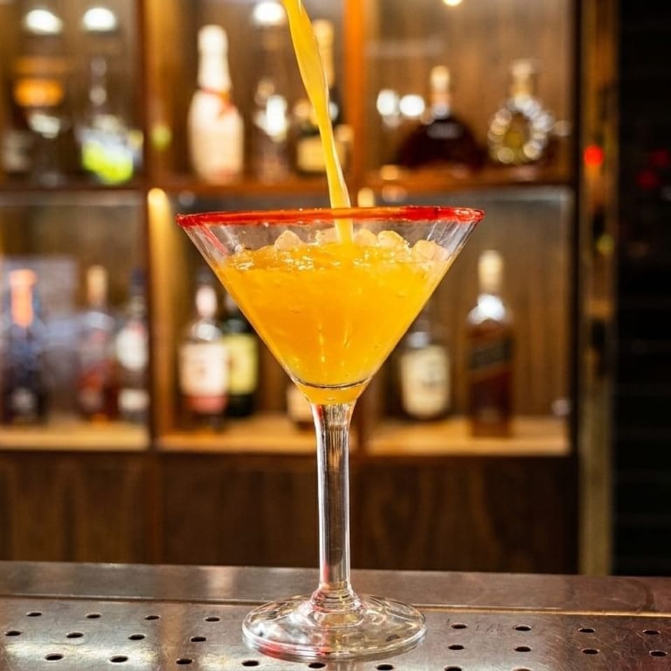 A bright orange cocktail with a red sugar rim is being poured into a martini glass at a bar.