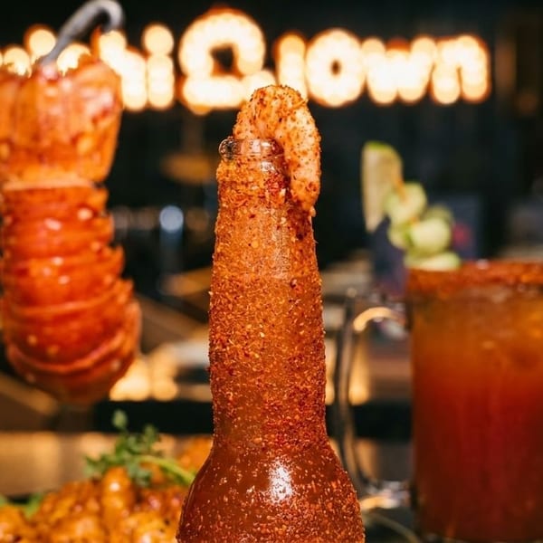 A spicy shrimp cocktail bottle and a lobster tail skewer on a dark restaurant table.